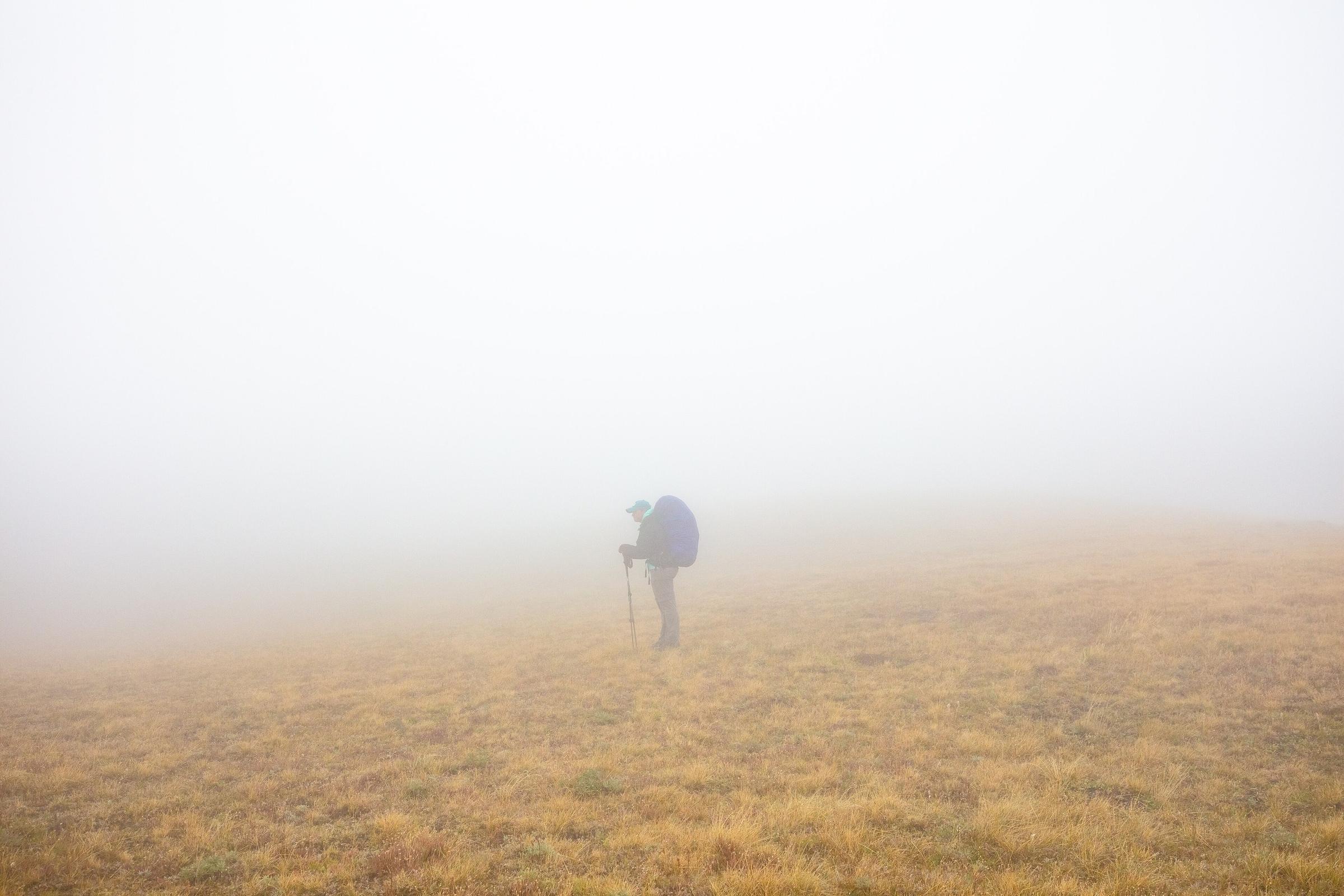 A backpacker stands in thick fog on an open meadow, with no trail in sight.