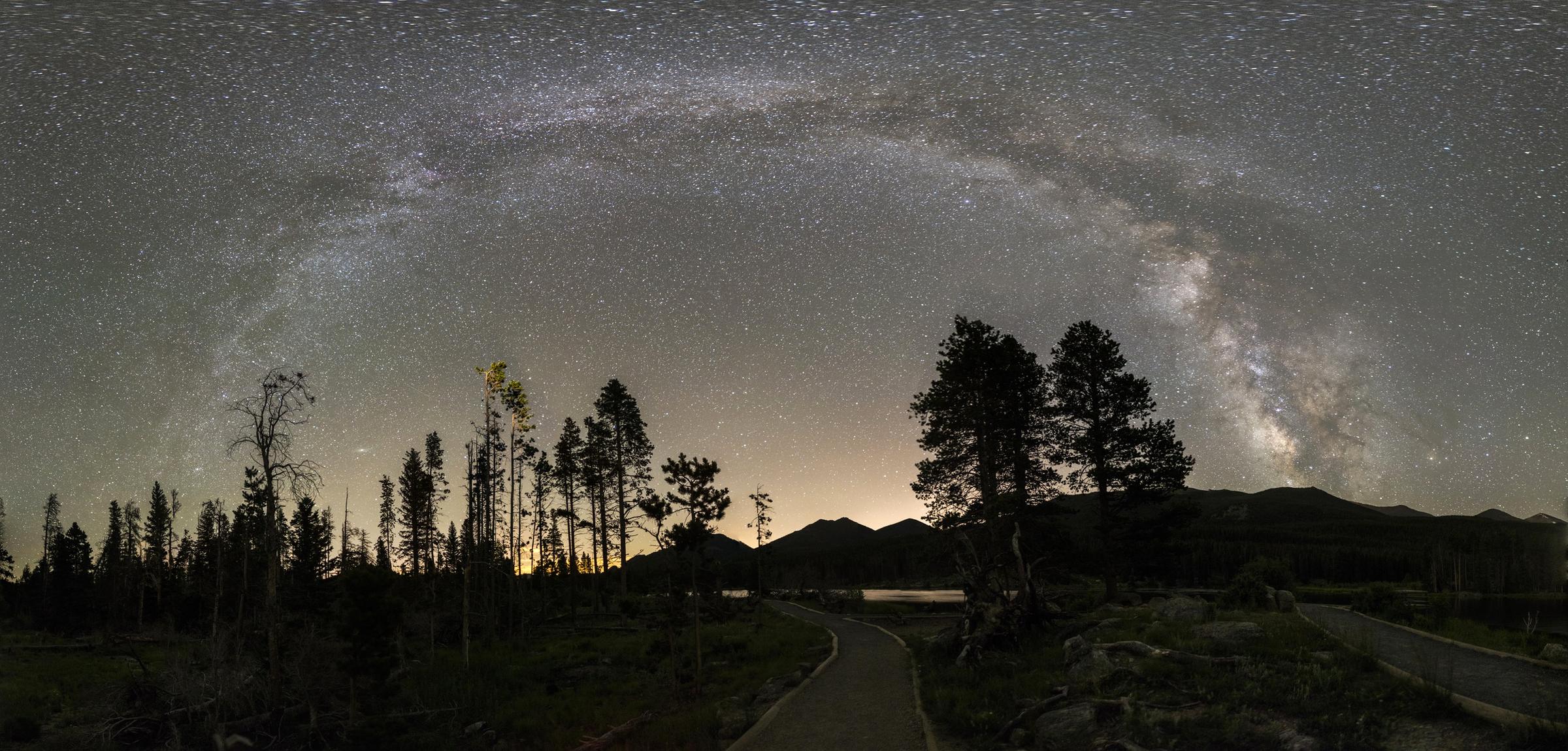 The Milky Way arches over pine trees and a path to a lake at night in Rocky Mountain National Park.