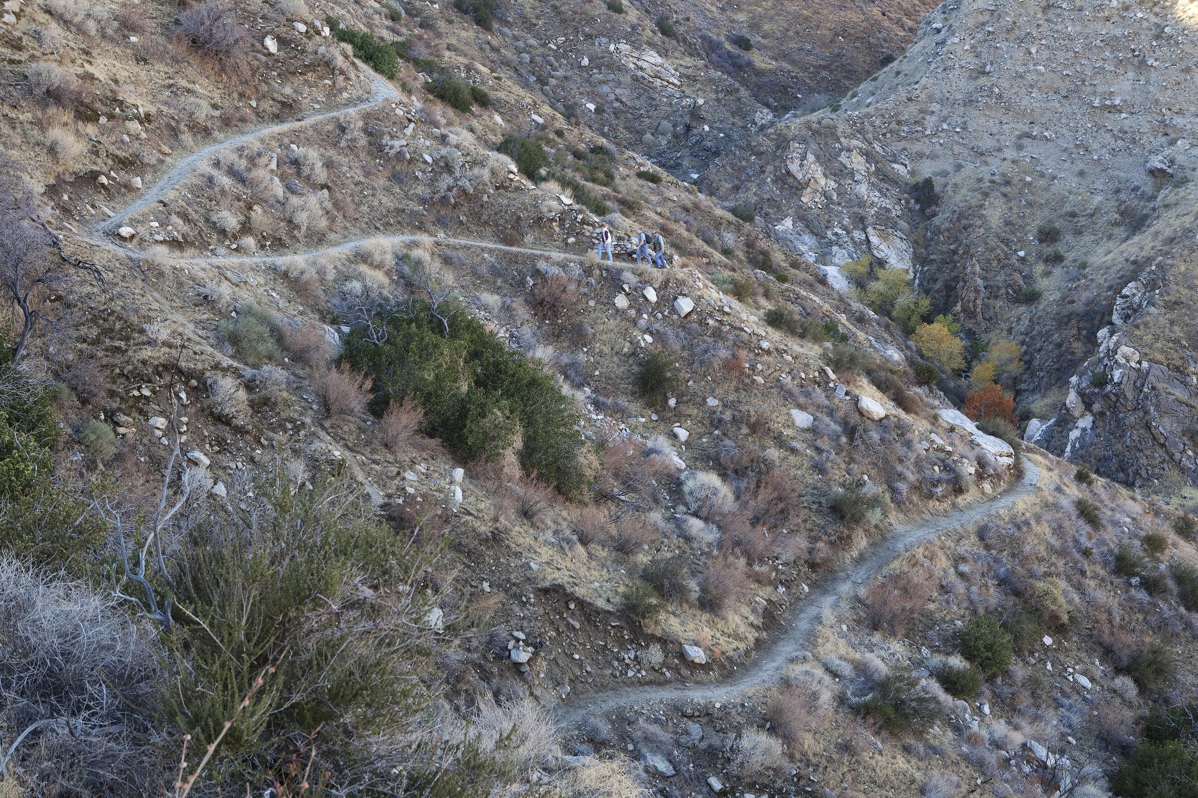 Three hikers on a switchback trail that zigzags up a dry, rocky canyon slope.